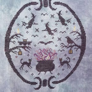 May include: A black and white cross-stitch design of a cauldron with purple and white flowers, surrounded by bats, owls, and cats. The design is framed by a black oval with a crescent moon design at the top and bottom.