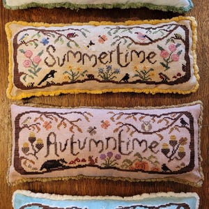 Puede incluir: Cuatro cojines rectangulares de punto de cruz con temas estacionales. El cojín superior es blanco con el texto Springtime, el segundo es beige con Summertime, el tercero es rosa con Autumntime y el inferior es azul claro con Wintertime.