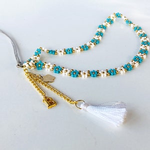 Puede incluir: Un collar de cuentas blancas y turquesas con una cadena dorada y un borla. El collar tiene un pequeño dije dorado con la letra "B" en él.