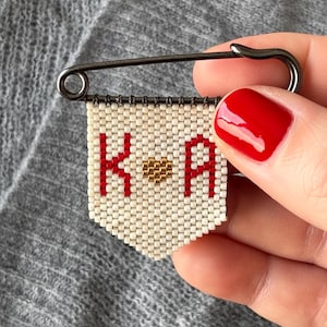 May include: A beaded banner pin featuring the letters "K" and "A" in red, with a gold heart between them, set against a white backdrop. The pin is attached to a silver safety pin.