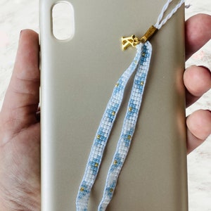 May include: A gold charm with a small flower design hangs from a white and blue beaded phone strap. The strap is attached to a gold phone case.