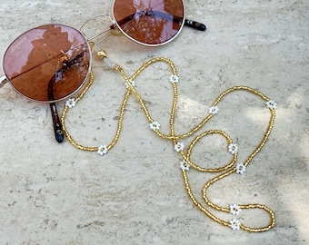 May include: Gold beaded glasses chain with white daisy accents. The chain is looped around a pair of brown sunglasses.
