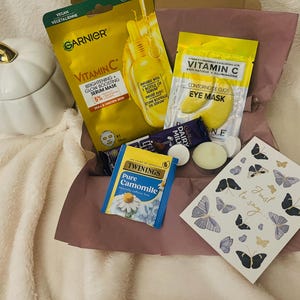 May include: A gift box containing a Garnier Vitamin C serum mask, a Vitamin C eye mask, a Cadbury Dairy Milk chocolate bar, a Twinings Pure Camomile tea bag, two small round candles, and a white greeting card with butterfly illustrations.