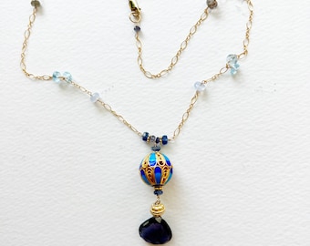 Puede incluir: Un collar de cadena dorada con detalles de piedras preciosas azules y transparentes. El colgante presenta un diseño de esmalte azul y dorado con una gota de piedra preciosa azul oscuro. El collar está sobre un fondo blanco.