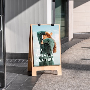 Peut inclure: Un panneau A en bois avec une affiche repr&eacute;sentant une femme portant un pull vert. L'affiche indique "It's Sweater Weather".