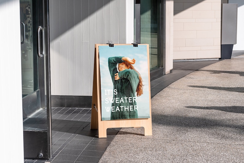 Peut inclure: Un panneau A en bois avec une affiche repr&eacute;sentant une femme portant un pull vert. L'affiche indique "It's Sweater Weather".