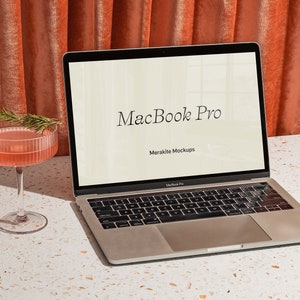 Op de afbeelding: Een zilveren MacBook Pro laptop met een wit scherm waarop de tekst "MacBook Pro" en "Merakite Mockups" wordt weergegeven. De laptop staat op een wit gespikkeld oppervlak met een roze cocktailglas op de voorgrond.