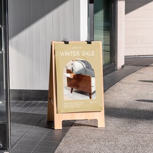 Peut inclure: Un panneau en bois brun en forme de A avec une affiche beige qui dit "Winter Sale" et pr&eacute;sente une photo d'une personne assise dans un fauteuil brun. Le panneau est devant un magasin avec une porte en verre.