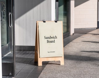 May include: A wooden A-frame sandwich board sign with a white background and black text that reads "Sandwich Board". The sign is standing on a pavement in front of a building.