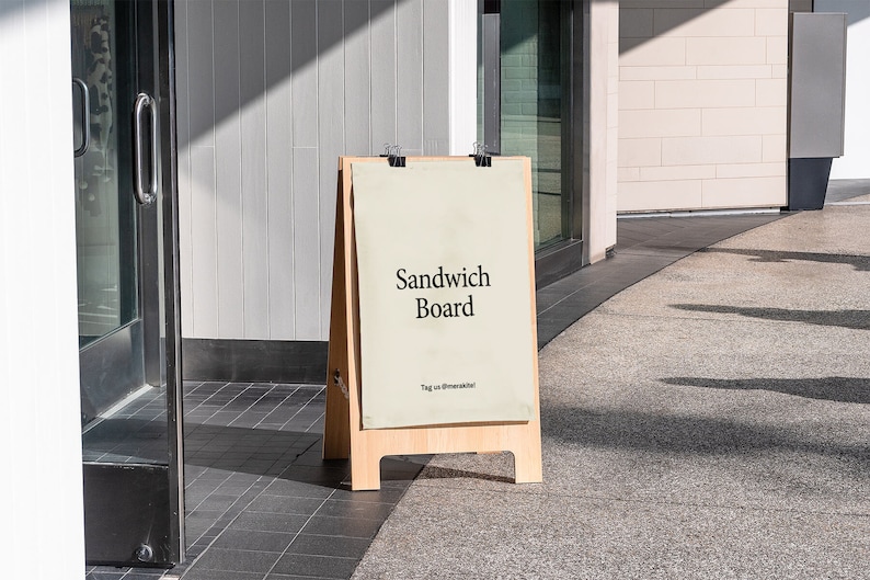Peut inclure: Un panneau d'affichage en bois en forme de A avec un fond blanc et du texte noir qui dit "Sandwich Board". Le panneau est debout sur un trottoir devant un b&acirc;timent.