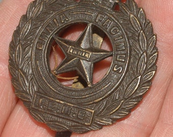 Puede incluir: Un distintivo de gorra militar de bronce con una estrella de cinco puntas, una corona y las palabras "Ubique Facimus" y "Je Me Souviens" alrededor de la estrella.