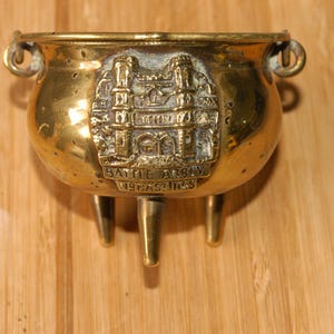 May include: A small, antique brass cauldron with three legs and two handles. The cauldron features an embossed design of a building and the text "BATTLE ABBEY HASTINGS". The cauldron is on a wooden surface.
