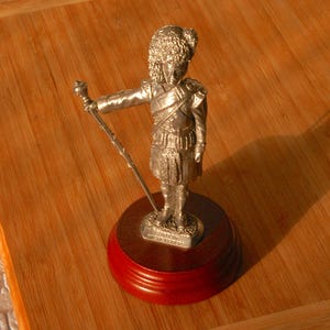 May include: A silver figurine of a Scottish Highlander in traditional dress, including a feathered bonnet and kilt. The figurine holds a staff and stands on a dark wooden base with a red rim. The base has text that reads "GORDON HIGHLANDERS".