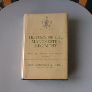 May include: A well-preserved book, "History of the Manchester Regiment", with black text on a tan cover. The title is prominent, with details of the First and Second Battalions from 1922-1948. Features a fleur-de-lis emblem.