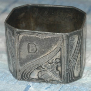 May include: Silver napkin ring with an ornate design featuring a stylised leaf and a letter 'D'.
