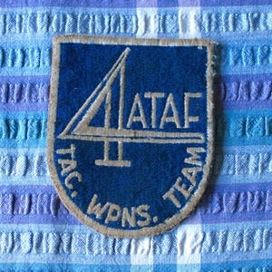 May include: A blue and tan patch with the text "4 ATAF II TAC WPNS. TEAM". The patch has a shield shape with a tan border and a design of a stylized number 4 and a horizontal line.
