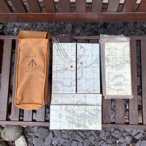 May include: A tan canvas carrying case labeled Millward Storecraft 40 MOD next to a set of folding map panels featuring grid lines and geographical terrain details, including a metal protractor tool attached to one of the map sections.