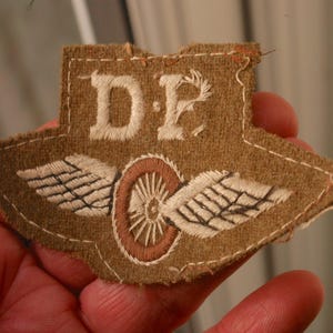 May include: A brown fabric patch with white embroidered wings and the letters "D.F." in white. A brown wheel is embroidered in the center of the patch.
