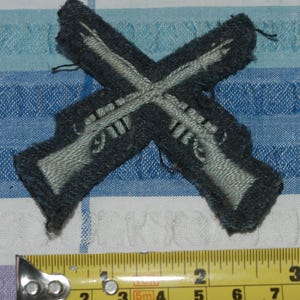 May include: A gray felt patch with two crossed rifles in white thread. The patch is approximately 2 inches wide.