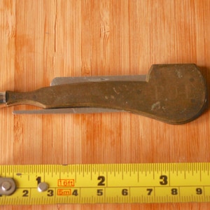 May include: A vintage brass pocket knife with a bottle opener and a small blade. The knife is approximately 10 cm long and rests on a wooden surface. A yellow measuring tape is in the foreground.