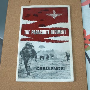 May include: A vintage print featuring the text "THE PARACHUTE REGIMENT" in white against a red background with parachute silhouettes. The image depicts soldiers in action with the word "CHALLENGE!" at the bottom.