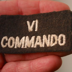 May include: A brown fabric patch with white embroidered text that reads "VI Commando".