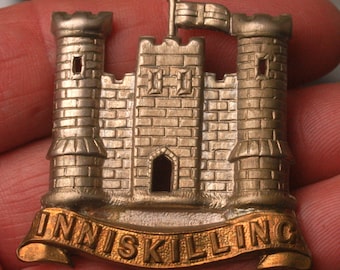 May include: A silver and gold metal badge with a castle design and a banner that reads "INNISKILLING".