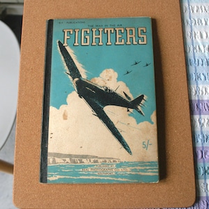 May include: A vintage book cover titled "FIGHTERS" with a teal background and an illustration of a fighter plane. The cover also features the text "THE WAR IN THE AIR" and the price "5/-". Published by Real Photographs Co. Ltd.