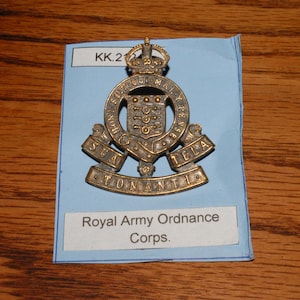 May include: A vintage Royal Army Ordnance Corps badge. The badge is made of a gold-colored metal and features a crown, a shield with keys, and the text "Royal Army Ordnance Corps." The badge is displayed on a blue paper with the text "KK.2".