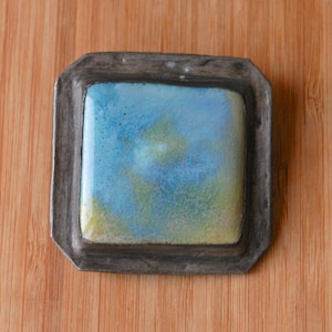 May include: A square, decorative dish with a blue, green, and yellow watercolor design. The dish is set in a dark gray metal frame. The background is a light brown wooden surface.