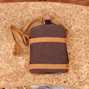 May include: A vintage, dark gray canteen with a tan canvas strap and accents. The canteen has a cork stopper and is wrapped with tan canvas bands. The canteen is designed to be worn over the shoulder.
