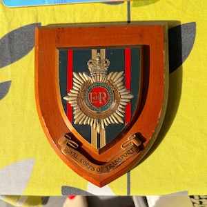 May include: A wooden wall plaque featuring the Royal Corps of Transport emblem. The shield-shaped mount displays a central crest with a crown, the letters ER, and a circular border with text. A banner at the bottom reads ROYAL CORPS OF TRANSPORT.