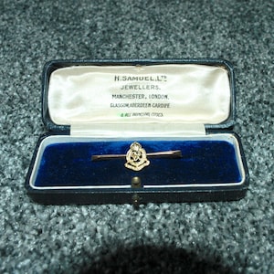 May include: A gold tie bar with an ornate emblem, displayed in an open, vintage black jewellery box with a blue velvet lining. The box lid features the text "H. Samuel Ltd. Jewellers, Manchester, London, Glasgow, Aberdeen, Cardiff & All Principal Cities."