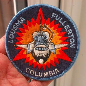 Op de afbeelding: Een blauwe en witte geborduurde patch met de tekst "LOUSMA FULLERTON COLUMBIA" rond een gestileerd beeld van een spaceshuttle met een rode en oranje explosie erachter.