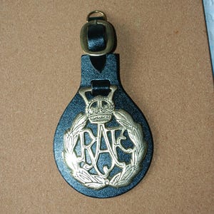 May include: A black leather key fob with a brass-colored RAF emblem. The emblem features a crown above the letters "RAF" within a laurel wreath. The fob has a brass-colored buckle and a leather loop for attachment.