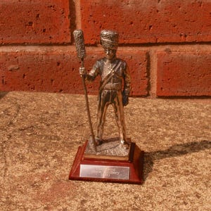 May include: A silver-colored figurine of a soldier in a uniform, holding a staff with a decorative top. The figurine stands on a dark wooden base with a silver plaque that reads "The Royal Horse Artillery 1840". The background is a red brick wall.