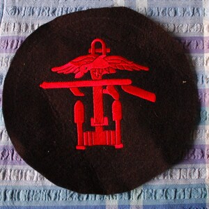 May include: A dark brown beret with a red embroidered emblem. The emblem features a stylized design with wings, a horizontal bar, and vertical elements. The beret is circular and rests on a checkered fabric background.