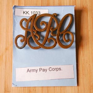 May include: A vintage brass emblem featuring the intertwined letters "APC" on a light blue card. The card also displays the text "Army Pay Corps." and the number "KK.1033". The emblem is set against a wooden background.