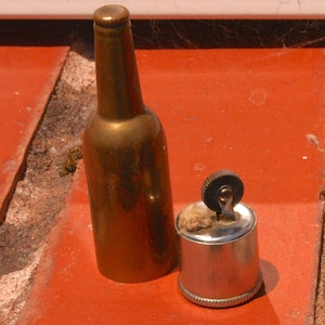 May include: A vintage brass bottle-shaped lighter and a small, cylindrical metal lighter with a wick. The bottle lighter is gold-toned, and the other lighter is silver-toned. Both are set against a red surface.