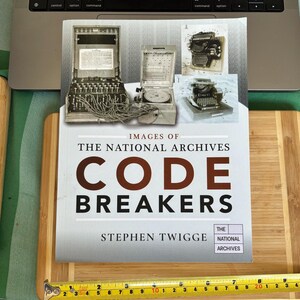 May include: A book titled "CODE BREAKERS" by Stephen Twigge, with images of historical code-breaking machines and typewriters. The book rests on a wooden board with a measuring tape. The National Archives logo is present.