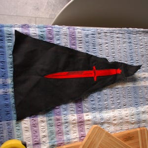 May include: A black pennant flag featuring a red sword emblem. The flag is triangular in shape and made of fabric. The red sword is centrally positioned, with a long blade and a cross-shaped hilt. The flag is displayed on a patterned surface.