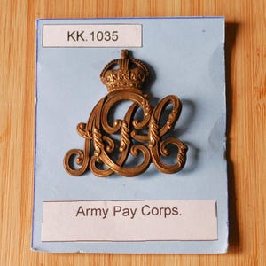 May include: A brass-colored Army Pay Corps badge with a crown on top and the letters "APC" intertwined. The badge is mounted on a light blue card with the text "KK.1035" at the top and "Army Pay Corps." at the bottom.
