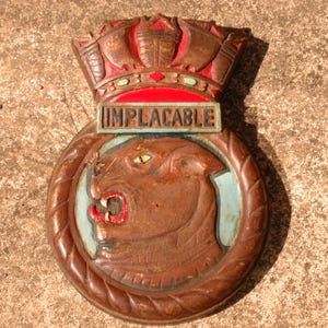 May include: A carved wooden plaque with a crown, the word "IMPLACABLE", and a roaring lion's head. The plaque is primarily brown with red, blue, and yellow accents. The lion has an open mouth with red teeth.