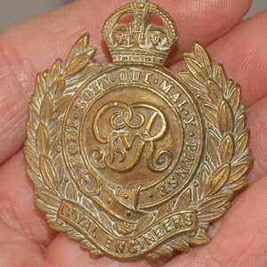May include: A gold-coloured metal badge with a wreath of leaves surrounding a crown and a royal crest. The text "HONI SOIT QUI MAL Y PENSE" and "ROYAL ENGINEERS" are inscribed on the badge.