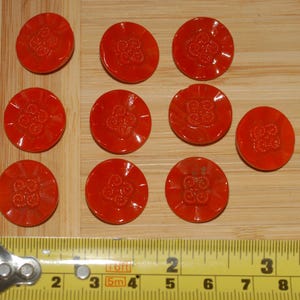 May include: Ten round, bright orange buttons with a floral design are displayed. The buttons are on a light brown wooden surface next to a yellow measuring tape. Each button is approximately 2.5 cm in diameter.