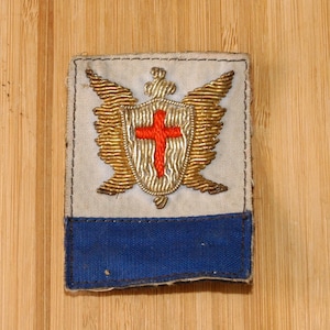 Op de afbeelding: Een rechthoekige patch met een witte achtergrond, met een goudkleurig schild met een rood kruis en gouden vleugels. Het onderste gedeelte is blauw. De patch is waarschijnlijk een embleem of insigne.