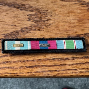 May include: A military ribbon bar featuring three distinct sections mounted on a black felt backing. The left ribbon is light blue with white stripes and a gold bar labeled LIBAN. The center is magenta with blue stripes, and the right is light blue with green and white stripes.