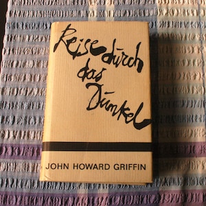 May include: A vintage book titled "Reise durch das Dunkel" by John Howard Griffin. The book has a tan cover with black lettering and a black horizontal stripe. The book is resting on a patterned fabric.