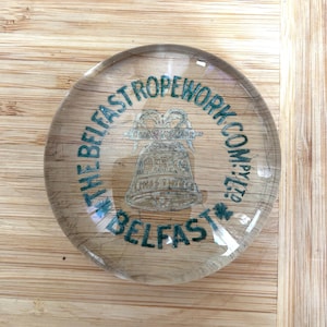May include: A clear, round paperweight with the text "THE BELFAST ROPEWORK COMPANY" in teal. The paperweight features a bell graphic and the word "BELFAST". The paperweight is on a wooden surface.