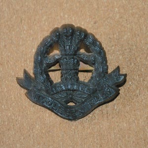 May include: A dark grey metal military badge. The badge features a crown, a wreath, and the words "MIDDLESEX REG." and "RUHER". The badge has a pin on the back.